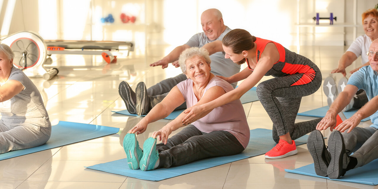 AdobeStock_341199351 Junger Trainer hilft älteren Menschen beim Training im Fitnessstudio.