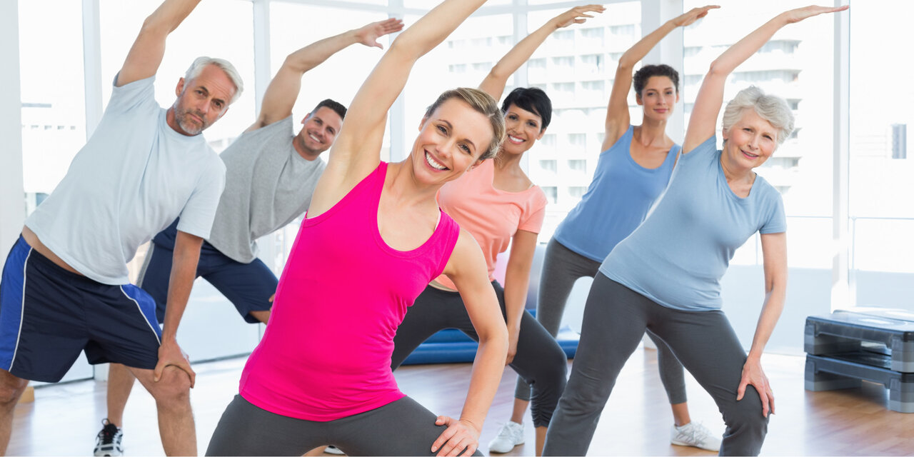 AdobeStock_63883845 Eine Gruppe an Menschen, die beim Yoga-Kurs die Hände streckt.